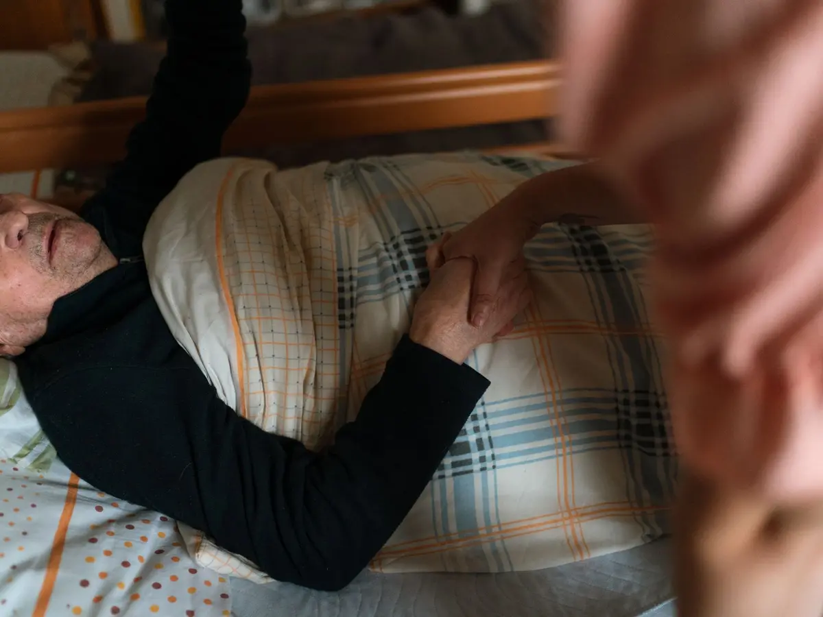 A caregiver holding an elderly woman’s hand as she rests in bed