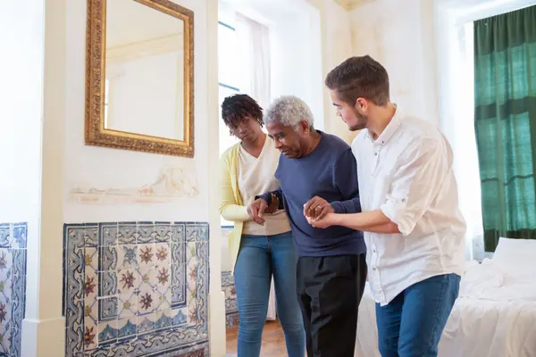 A caregiver supporting an elderly man as he walks across his living room