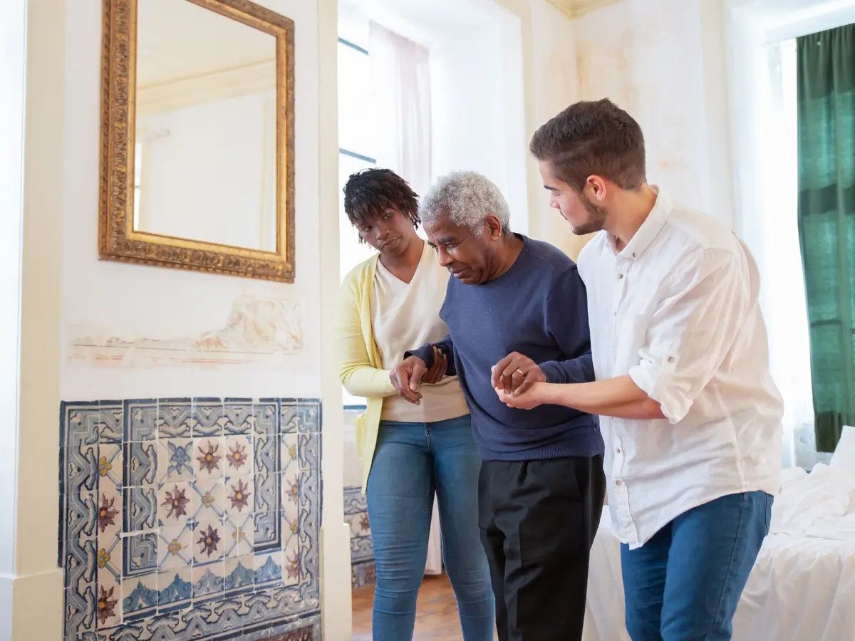 A caregiver supporting an elderly man as he walks across his living room
