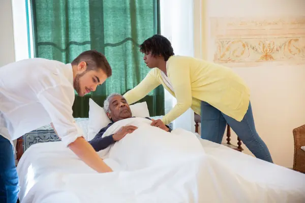 Two caregivers helping a man sit up comfortably in bed at home