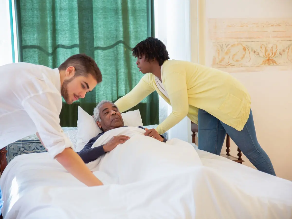 Two caregivers helping a man sit up comfortably in bed at home
